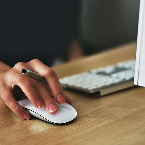 Nutzung digitaler Beschaffungssysteme Eine Nahaufnahme der Hand einer Person, die eine weiße Computermaus benutzt. Die Hand liegt auf einem Holzschreibtisch in der Nähe einer Tastatur und eines Computermonitors.