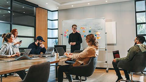 Eine Gruppe von Menschen hat in einem modernen Büro eine Besprechung. Eine Person steht an einem Whiteboard mit Haftnotizen, während die anderen an einem Tisch mit Laptops sitzen, zuhören und diskutieren.