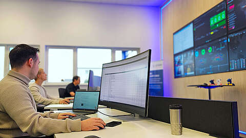 In einem modernen Büro sitzen mehrere Personen an Schreibtischen mit großen Computermonitoren. Eine Person tippt auf einer Tastatur, während große Wandbildschirme Daten und Diagramme anzeigen. Der Arbeitsbereich ist mit blauem Akzentlicht hell erleuchtet.