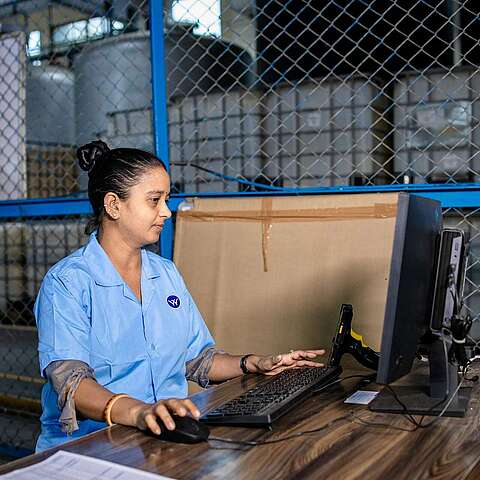 Bedarfsgerechte Planung und Strategie Eine Frau in blauer Uniform sitzt an einem Schreibtisch und arbeitet an einem Computer in einer Industrieanlage. Hinter ihr sind durch einen Metallzaun große Lagerbehälter sichtbar. Auf dem Schreibtisch liegen ein Ventilator und einige Dokumente.