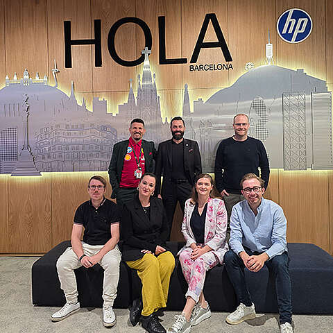 Sieben Personen stehen in einem Raum, lächeln und zeigen den Daumen nach oben vor einem Bildschirm, auf dem die Logos von „HP Amplify Elevate“ und verschiedenen Technologieunternehmen zu sehen sind.