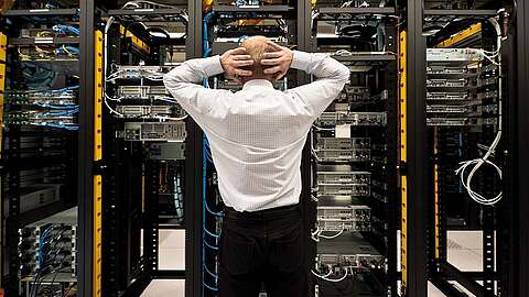 Eine Person steht mit den Händen auf dem Kopf vor einer Reihe von Server-Racks voller Netzwerkgeräte und Kabel in einem Rechenzentrum. Sie scheint die Server zu beobachten oder zu inspizieren.