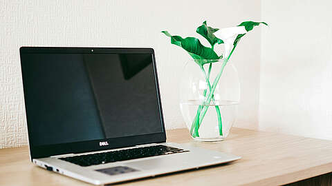 Hochwertige Laptops von Dell Ein geschlossener Dell-Laptop steht auf einem Holzschreibtisch neben einer durchsichtigen Glasvase mit grünen Blättern und weißen Calla-Lilien. Der Hintergrund ist eine helle, strukturierte Wand.