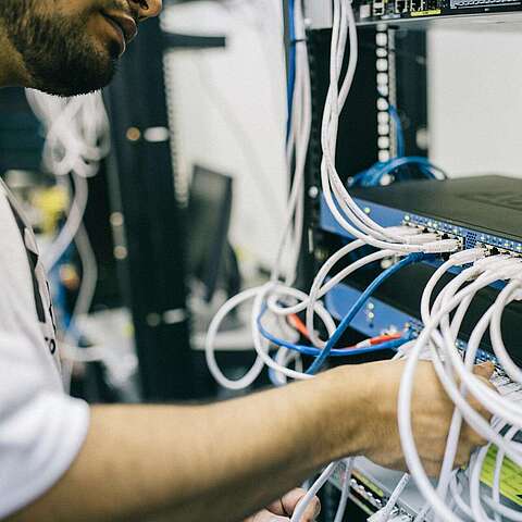 Nachhaltigkeit und Lebenszyklus-Management Eine Person in weißem Hemd verbindet oder organisiert mehrere Netzwerkkabel mit blauen Netzwerk-Switches, die auf einem Server-Rack montiert sind. Im Hintergrund sind weitere elektronische Geräte und Kabel zu sehen.