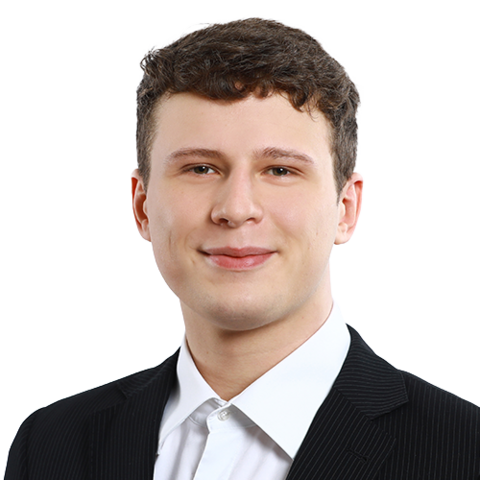 Ein junger Mann mit kurzen braunen Haaren, der eine schwarze Nadelstreifen-Anzugsjacke und ein weißes Hemd trägt, lächelt leicht in die Kamera vor einem schlichten weißen Hintergrund.