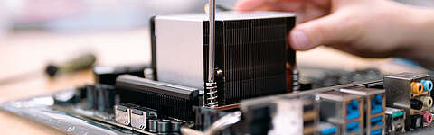 Nahaufnahme einer Hand, die einen großen Kühlkörper auf einer Computer-Hauptplatine installiert, wobei im Hintergrund verschiedene Anschlüsse und Komponenten zu sehen sind.