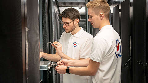 Zwei Männer in weißen Poloshirts mit einem Logo arbeiten gemeinsam an Geräten in einem Serverraum und konzentrieren sich dabei auf den Umgang mit Werkzeugen sowie die Überprüfung der Hardware im Inneren eines Serverracks.