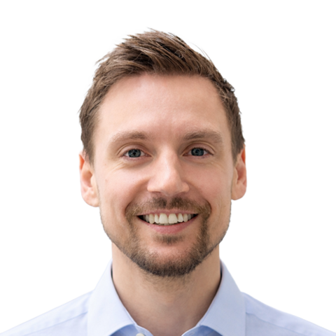 Ein lächelnder Mann mit kurzen hellbraunen Haaren und gestutztem Bart, der einen hellblauen Pullover trägt, wird vor einem schlichten weißen Hintergrund gezeigt.