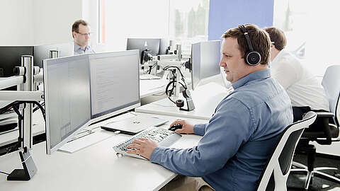 Eine Person mit Kopfhörern arbeitet an einem Schreibtisch mit zwei Monitoren. Mehrere andere sitzen an Schreibtischen in der Nähe und benutzen ebenfalls Computer. Das Büro ist hell und modern gestaltet, mit weißen Möbeln und natürlichem Licht.