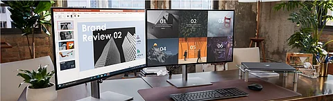 Auf zwei Monitoren auf einem Schreibtisch werden eine Präsentation und ein aus mehreren Feldern bestehendes Dashboard angezeigt. Auf dem Schreibtisch befinden sich eine Tastatur, eine Maus, ein Laptop, Pflanzen und Büromaterial. Im Hintergrund sind große Fenster und die Stadtlandschaft zu sehen.