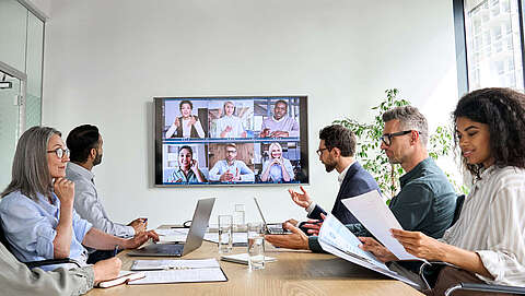 Mehrere Personen sitzen in einem Konferenzraum vor einem großen Bildschirm mit Arbeitsutensilien gemeinsam am Tisch