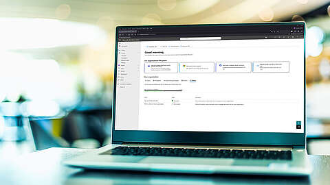 Ein Laptop mit dem Microsoft 365-Logo auf dem Bildschirm, mit einem unscharfen Hintergrund aus Lichtern und Tischen, der an ein Büro oder ein Café erinnert.