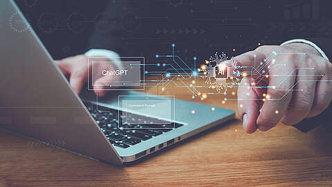 Eine Person zeigt auf eine Laptoptastatur, auf der digitale Grafiken mit den Symbolen "ChatGPT", "AI" und "Eingabeaufforderung" zu sehen sind, die künstliche Intelligenz und Technologieintegration symbolisieren.