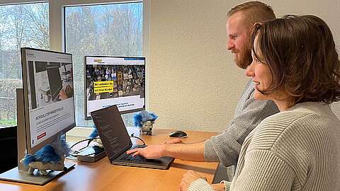 Zwei Personen arbeiten an einem Schreibtisch mit zwei Monitoren und einem Laptop. Einer tippt, während der andere zusieht. Auf dem Schreibtisch liegen kleine blaue und graue Stofftiere. Vor dem Fenster sind Bäume zu sehen.