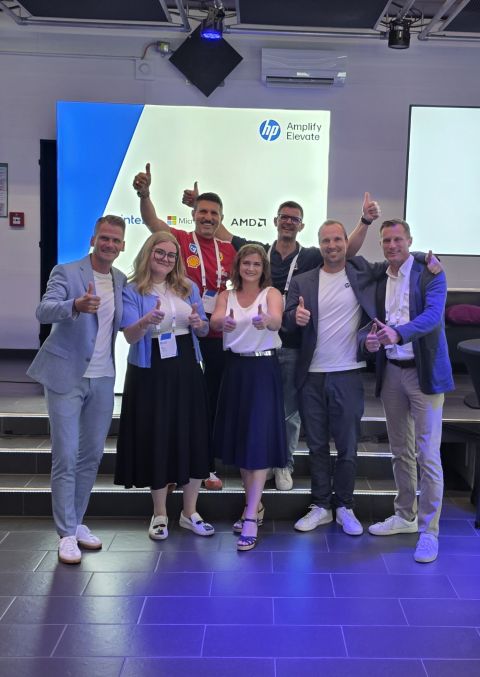 Sieben Personen stehen in einem Raum, lächeln und zeigen den Daumen nach oben vor einem Bildschirm, auf dem die Logos von „HP Amplify Elevate“ und verschiedenen Technologieunternehmen zu sehen sind.